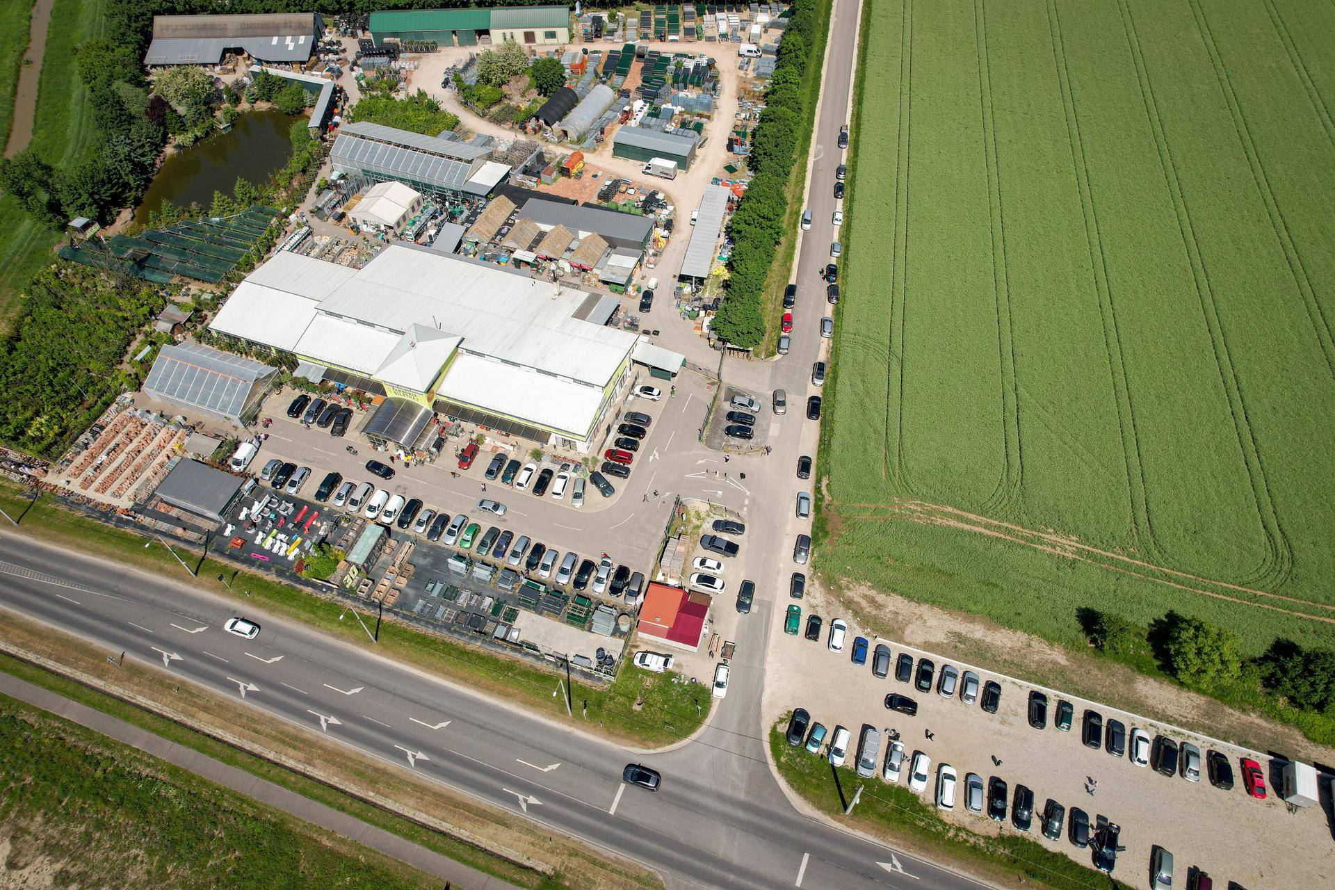 Luftaufnahme einer Gewerbefläche mit Parkplätzen, Straße und weiten grünen Feldern in der Umgebung.
