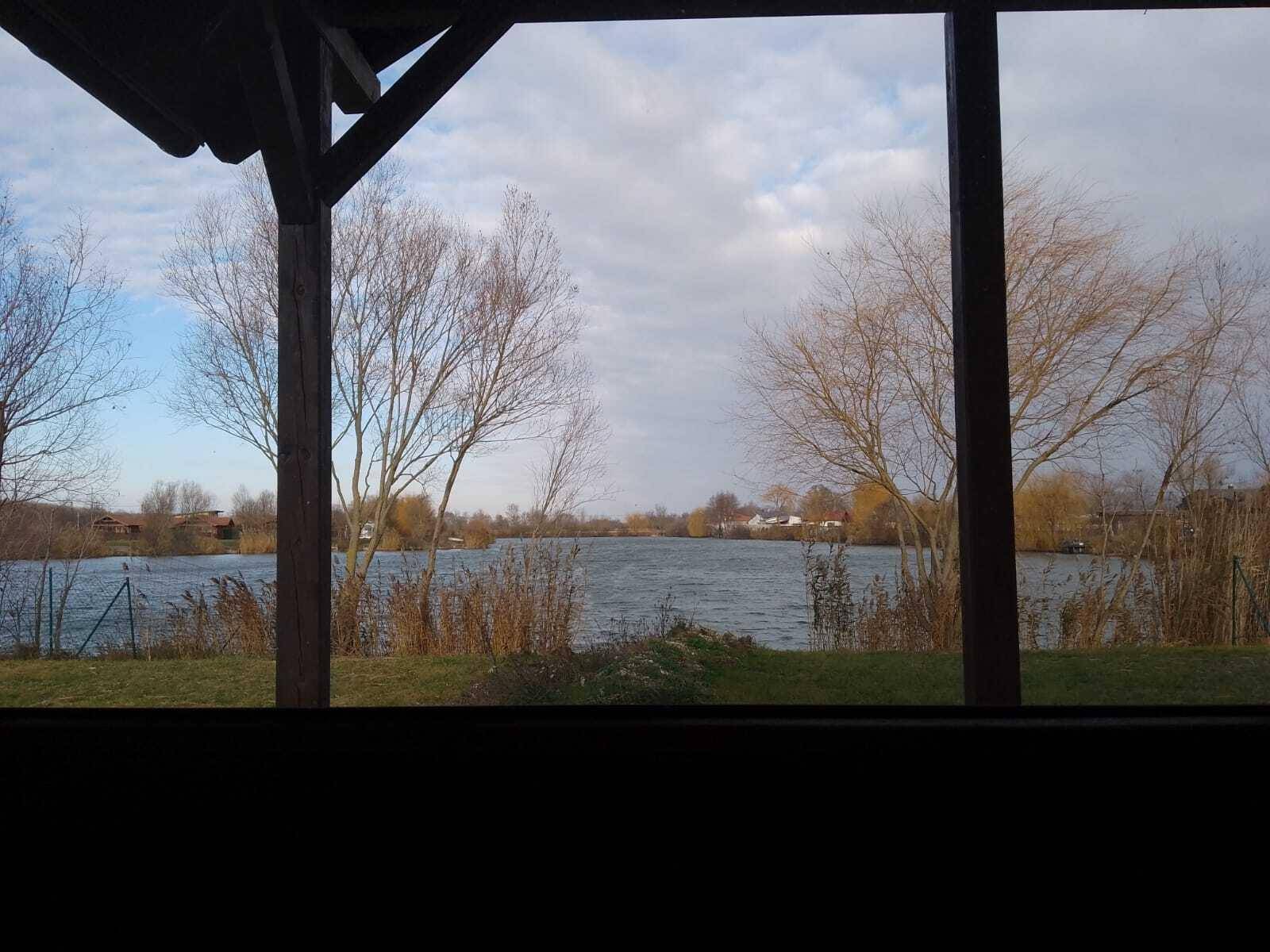 Blick von der überdachten Veranda auf den See mit Bäumen und Schilf am Ufer.