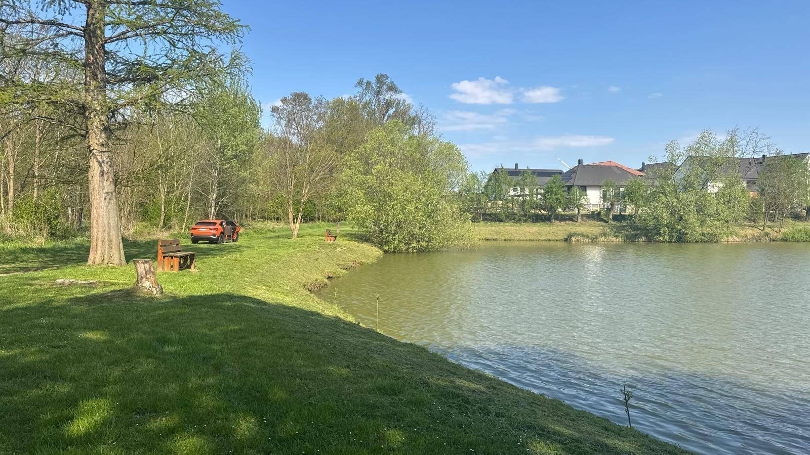 Idyllische Landschaft mit einem See, Bäumen, einer Bank und einem orangefarbenen Auto am Ufer.
