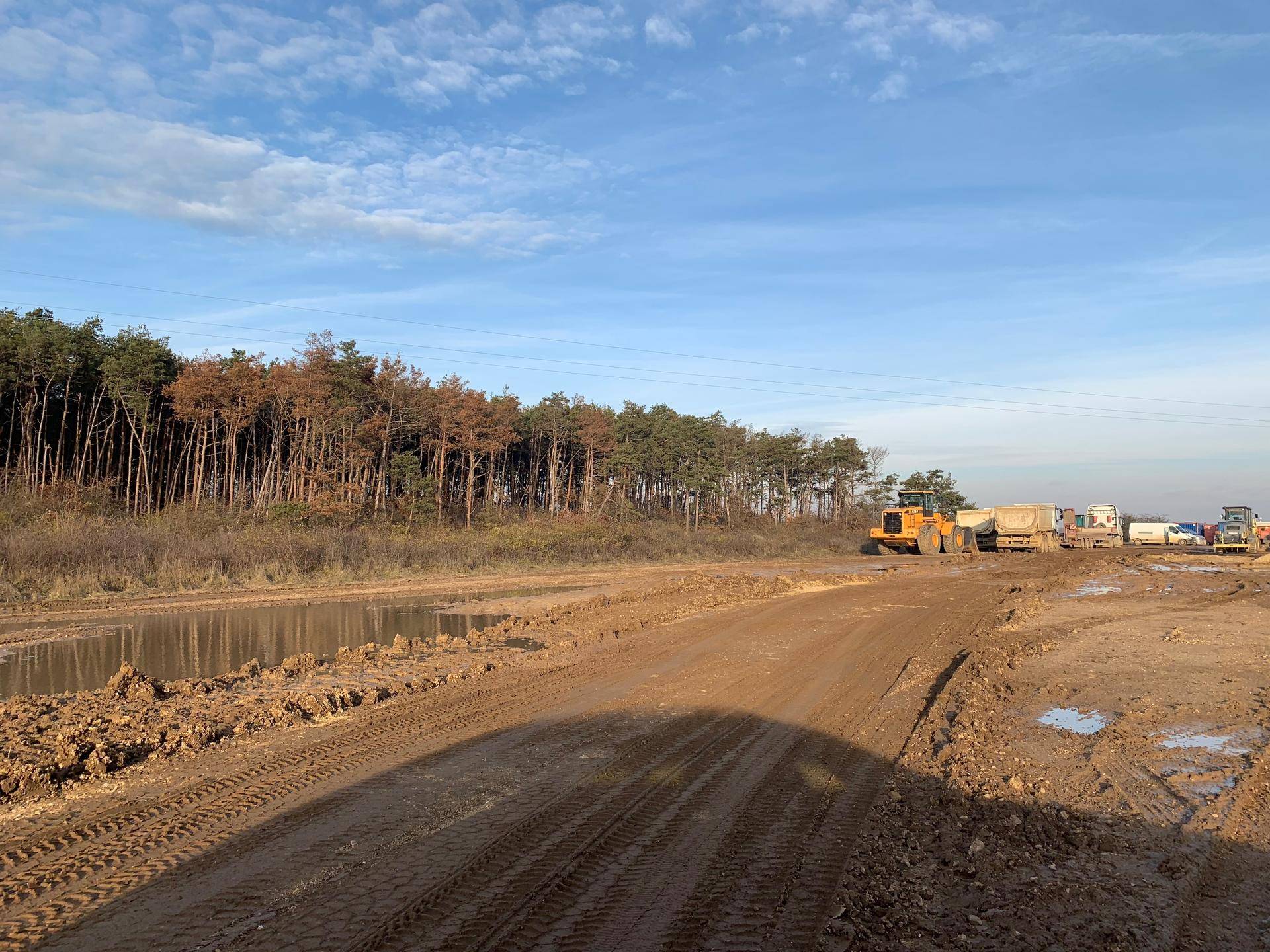Breiter, schlammiger Bauweg neben einem Waldgebiet mit Baumaschinen in der Ferne.