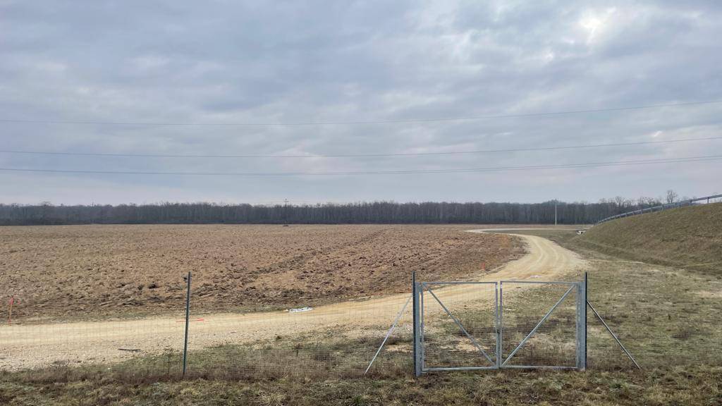 Weites Feld mit einem Feldweg und einem Metalltor unter einem bewölkten Himmel.