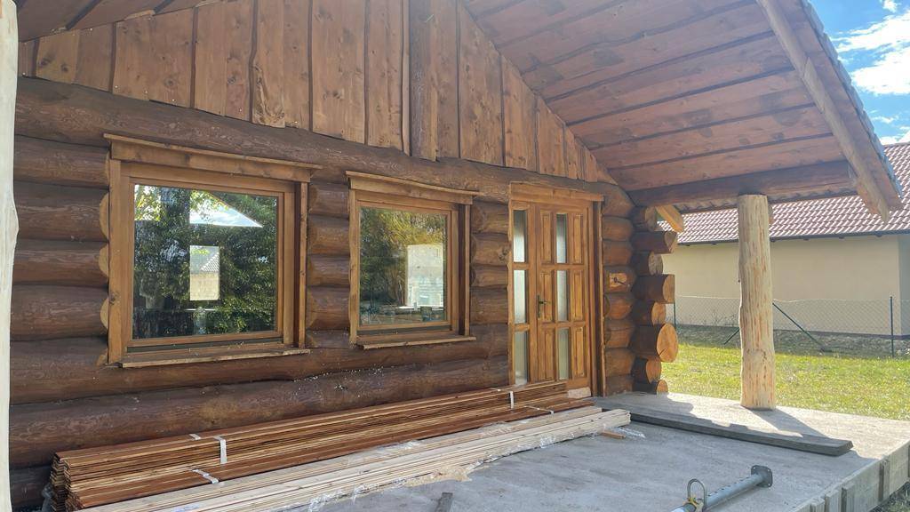 Blockhaus-Eingangsbereich mit Holztür, Fenstern und gestapelten Holzbrettern auf der Terrasse im Sonnenlicht.