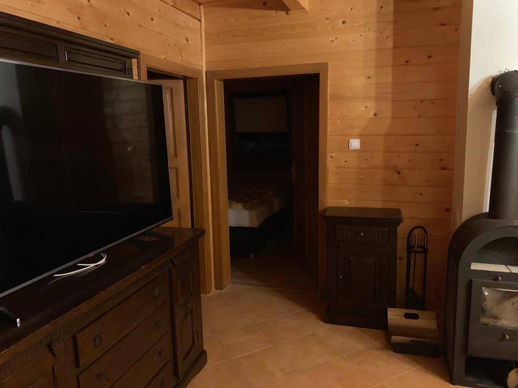 Wohnbereich mit Fernseher, Holzmöbeln und Zugang zu einem weiteren Zimmer, inklusive Kaminofen.