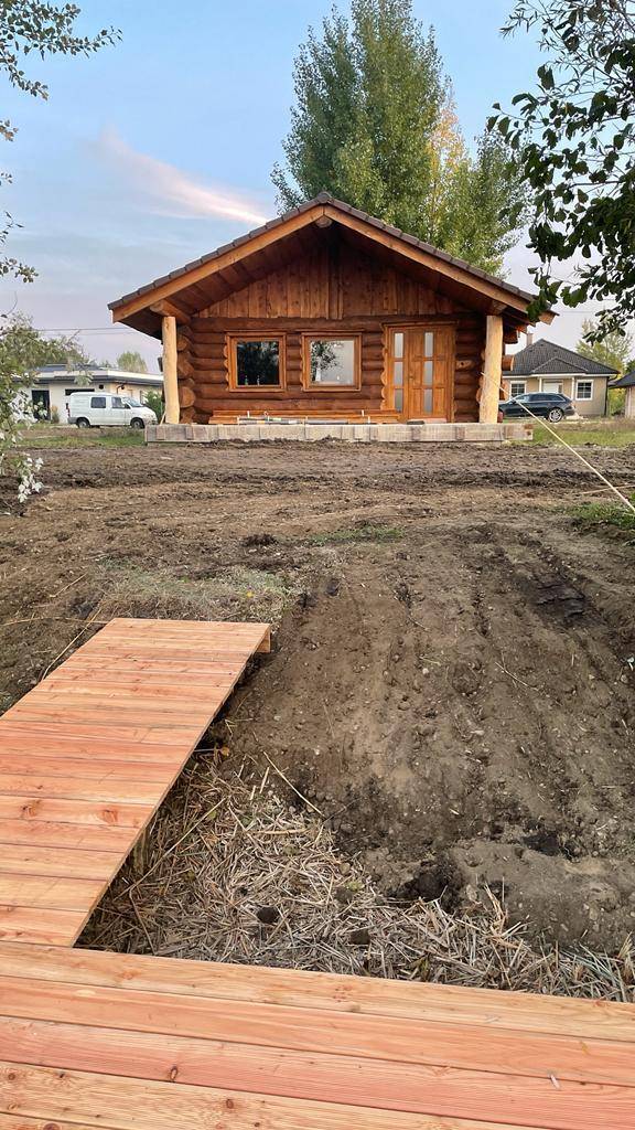 Blockhaus mit Holzsteg über unebenem Gelände, umgeben von ländlicher Natur und Bäumen.