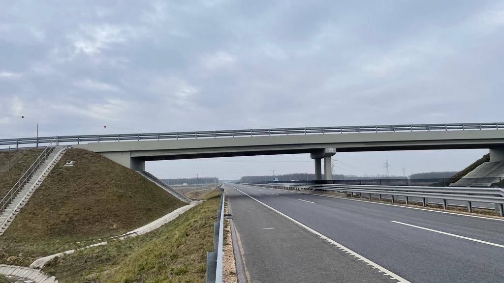 Autobahn unter einer Brücke mit weiten Feldern und bewölktem Himmel in der Ferne.