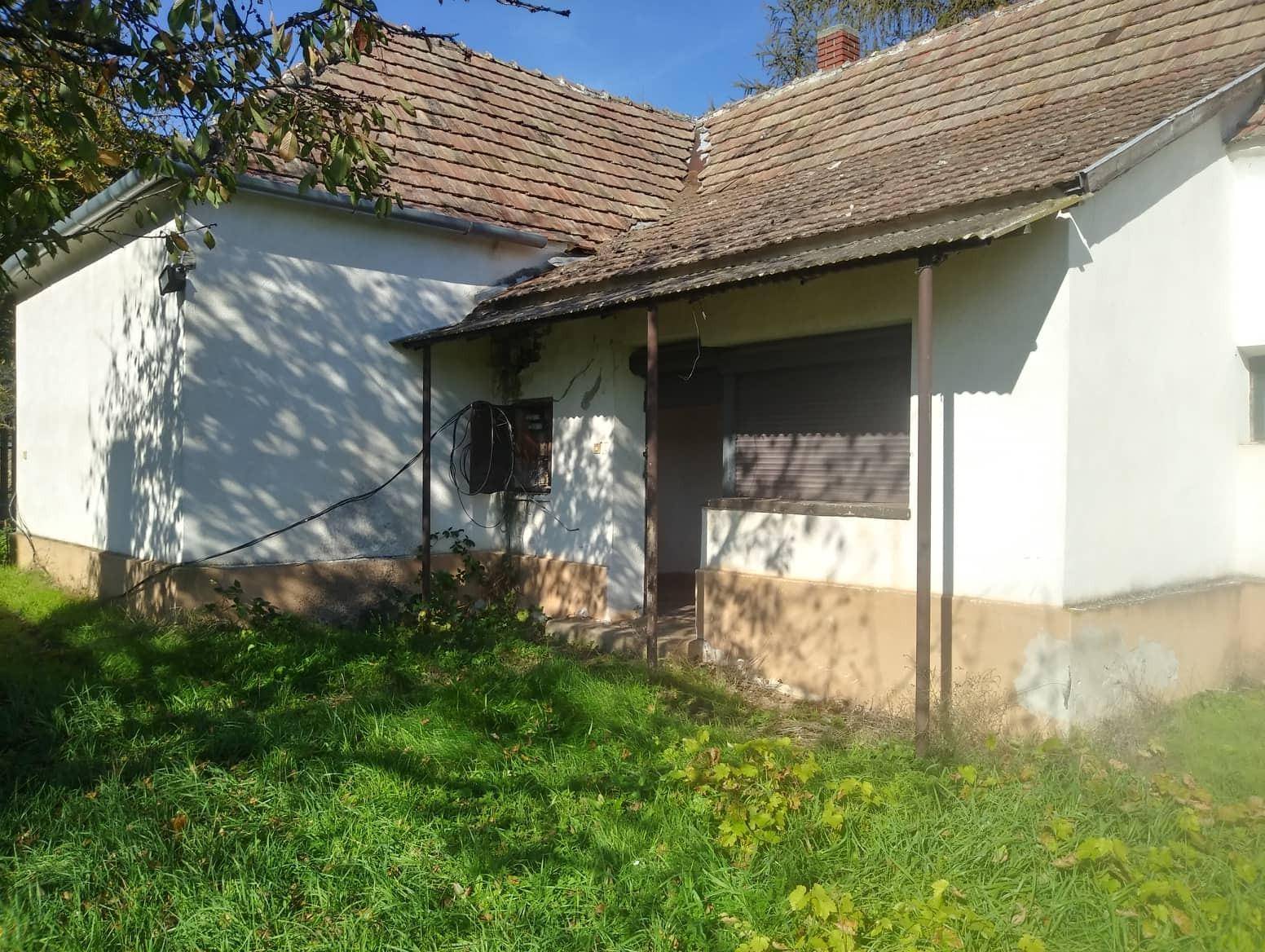 Seitenansicht eines älteren Hauses mit Vordach und einfacher Fassade, umgeben von Gras.