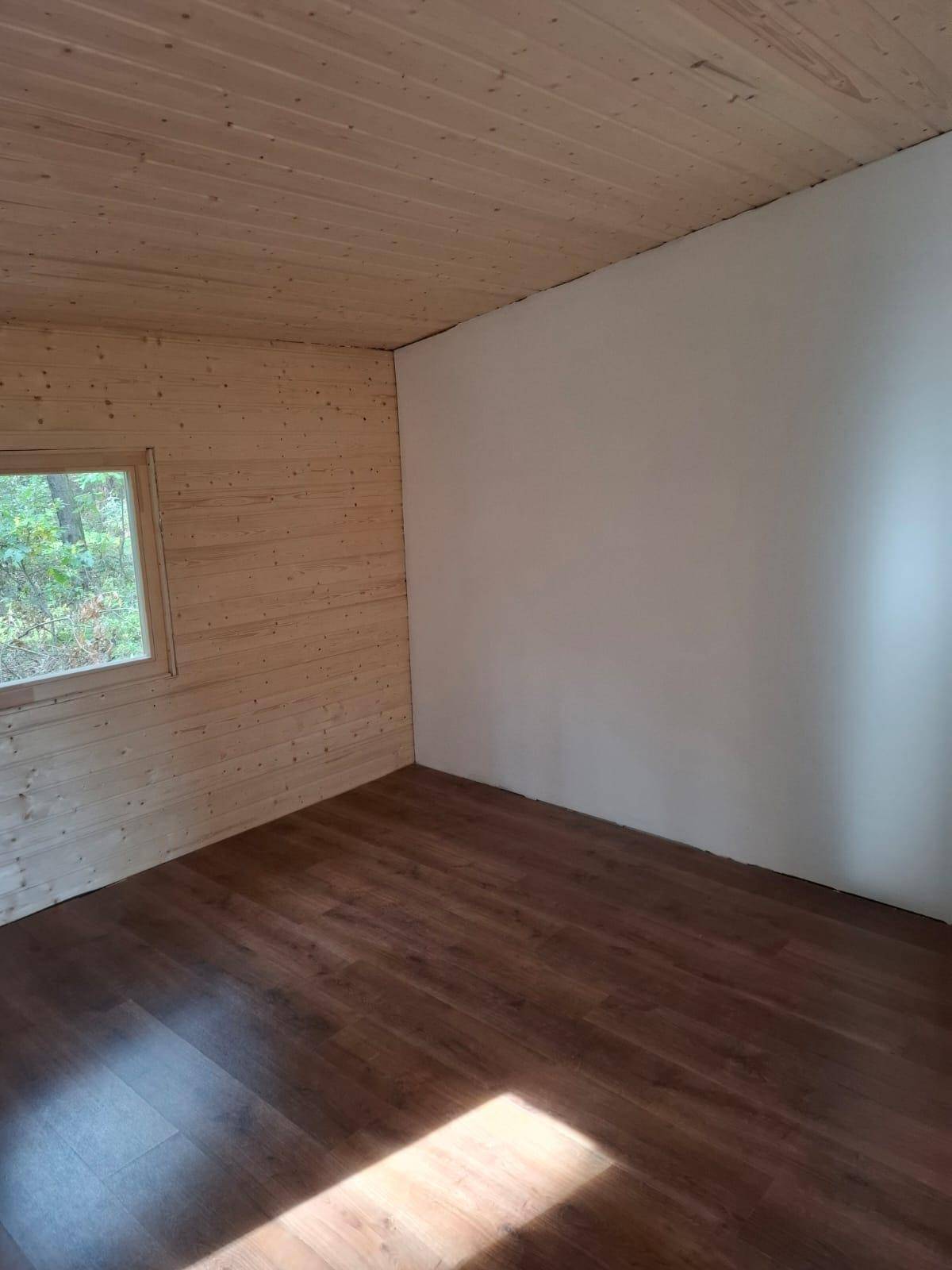 Schlichter Raum mit Holzwand, weißer Wand und Laminatboden, mit einem Fenster zum Wald.