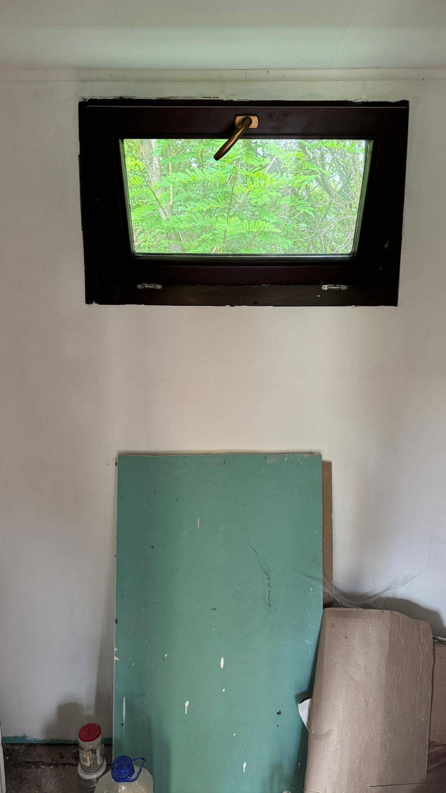 Kleines Fenster in einem Raum, das einen Blick auf dichtes Grün außerhalb des Hauses bietet.