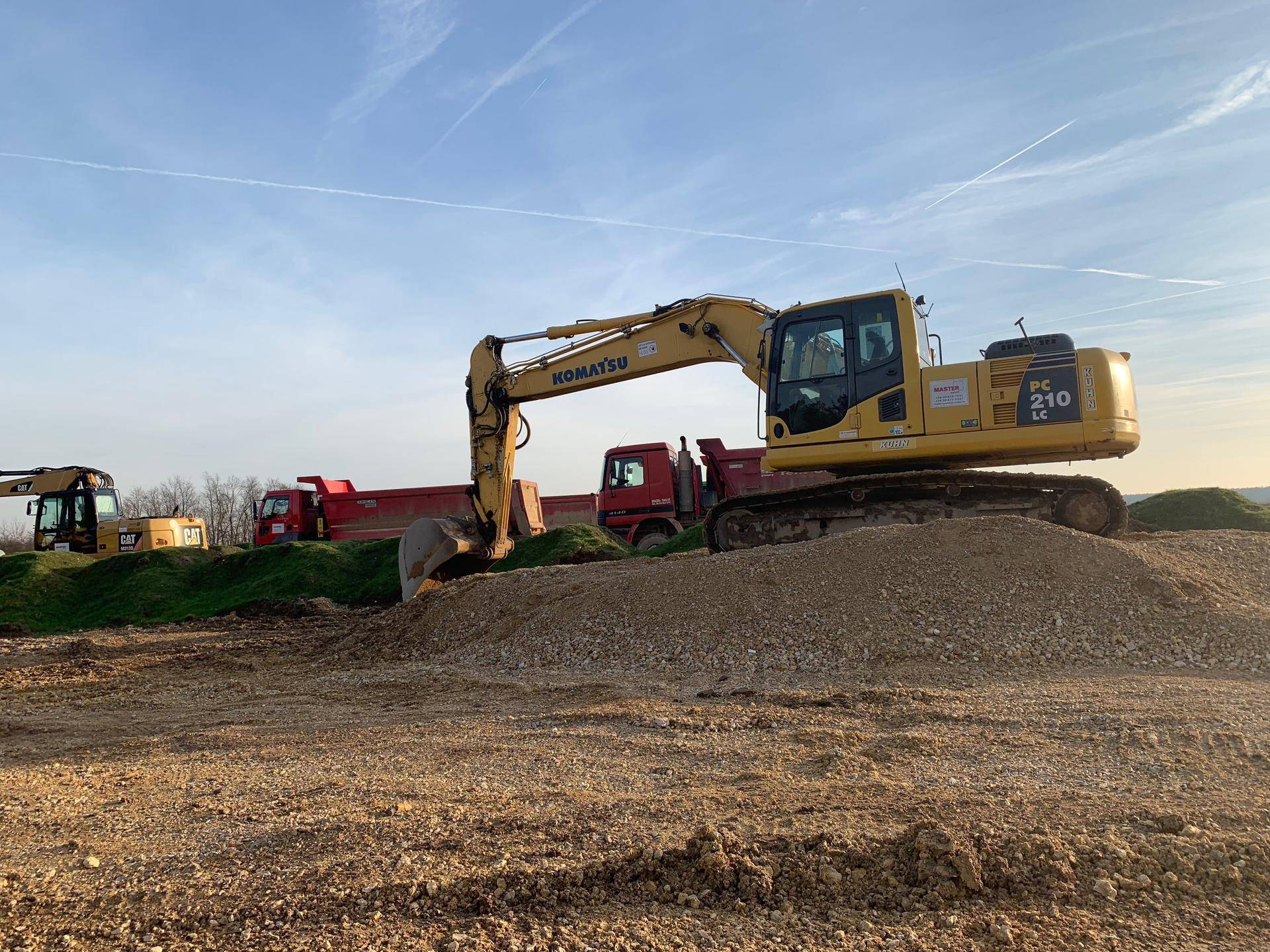Gelber Bagger auf einem Kieshaufen auf einer Baustelle mit roten Lastwagen im Hintergrund.