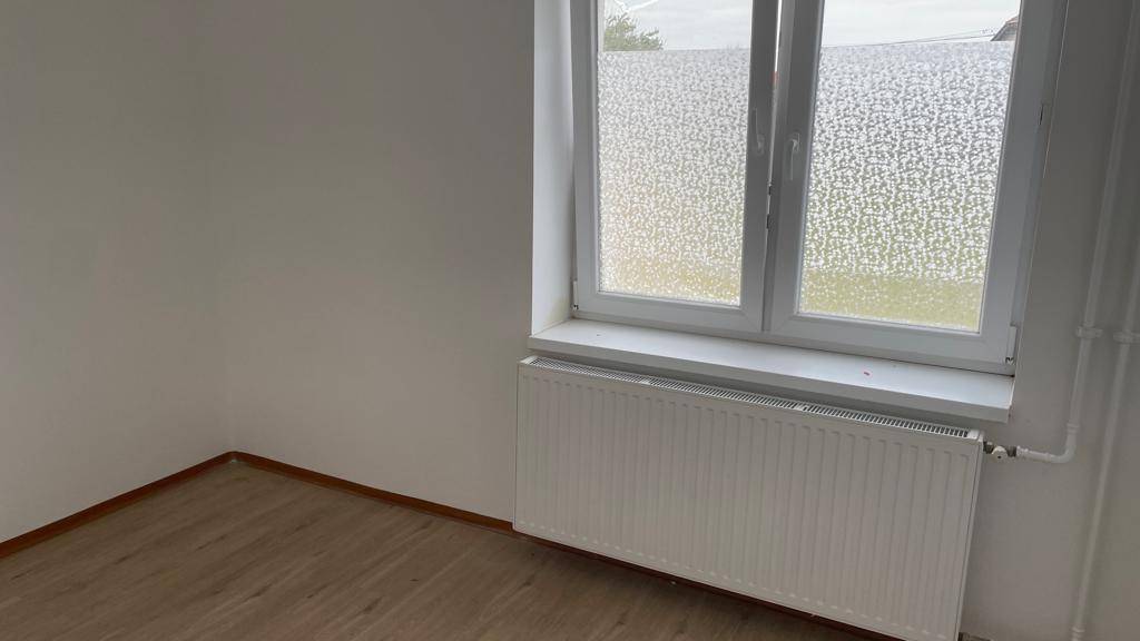 Heller Raum mit Laminatboden, weiß gestrichenen Wänden und einem großen Fenster mit Heizkörper.