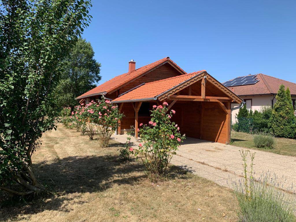 Holzhaus mit Carport und blühenden Rosenbüschen im Vorgarten, umgeben von Grün.