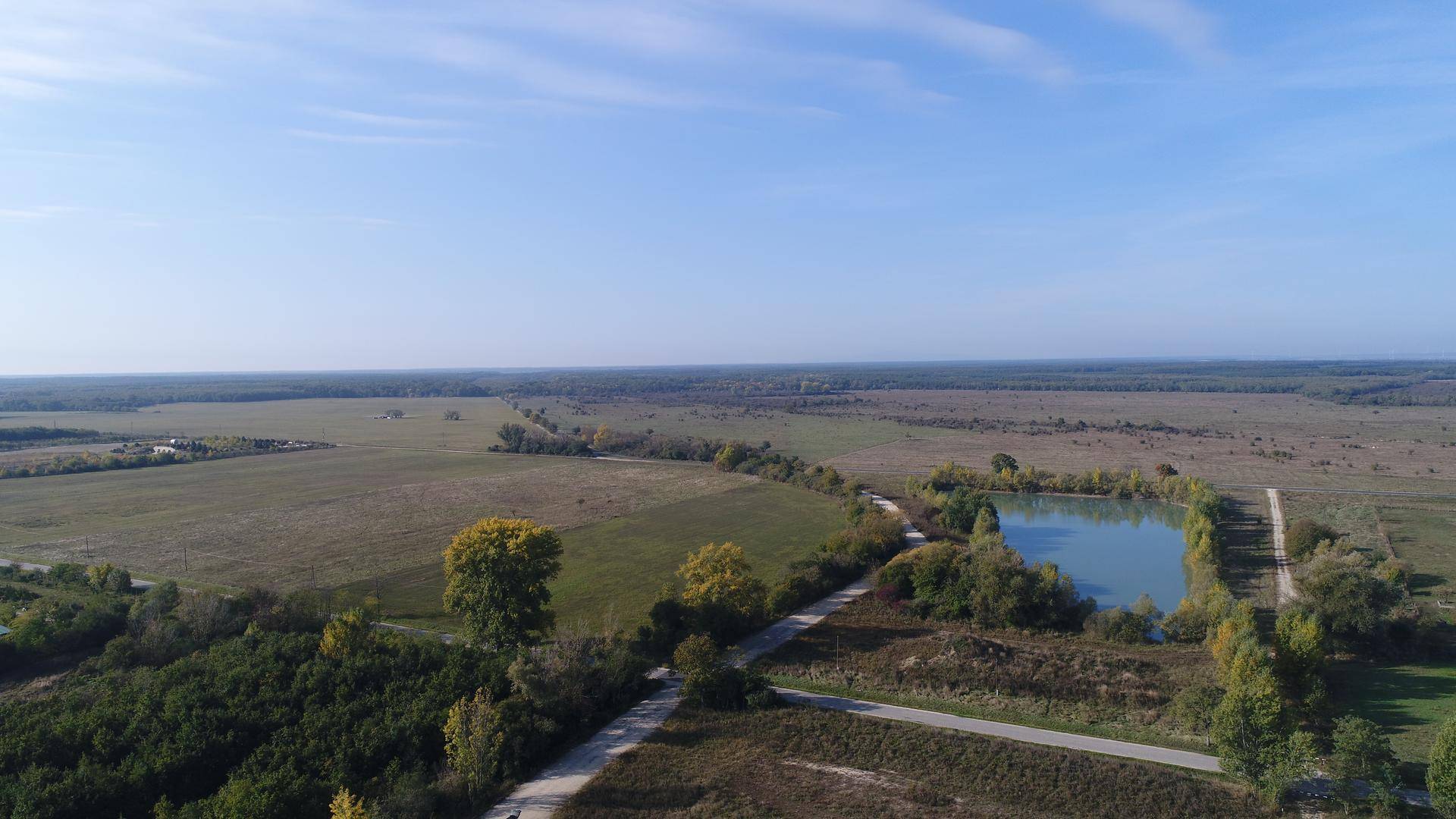Weitläufige Landschaft mit Feldern, Wäldern und einem Teich unter klarem Himmel, aus der Vogelperspektive.