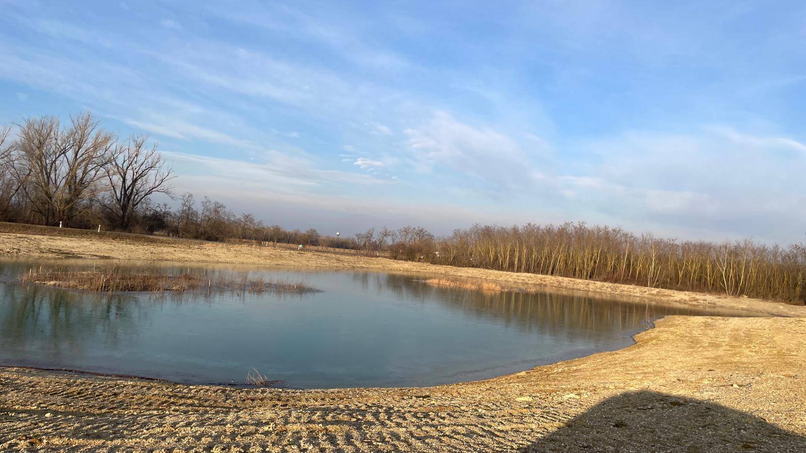 Weitläufiger See oder Teich mit kahlen Bäumen am Horizont und sandigen Ufern unter weitem Himmel.