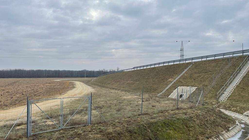 Böschung der Autobahn neben einem Feldweg und einem Metalltor mit Stromleitungen.