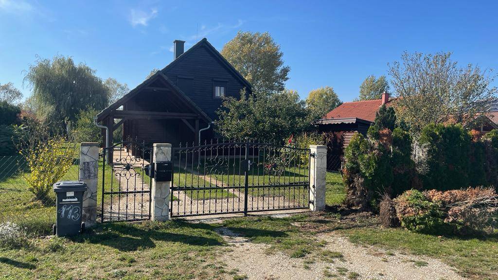 Eingangsbereich des Anwesens mit schmiedeeisernem Tor, Steinsäulen und Holzhaus im Hintergrund.