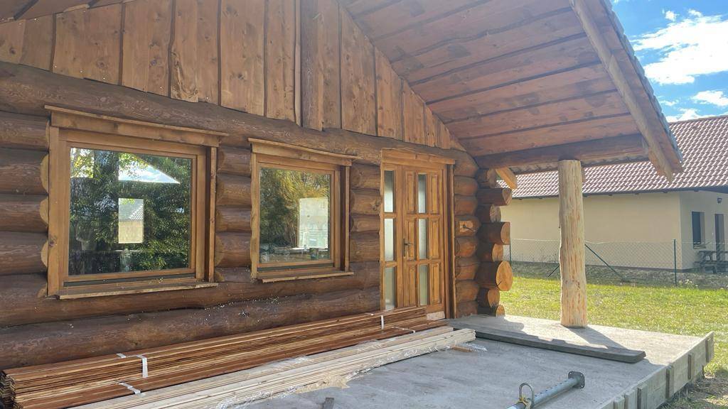 Vorderansicht des Blockhauses mit überdachtem Eingang und gestapelten Holzbrettern auf der Terrasse.