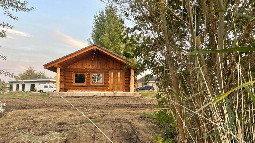 Blockhaus in Bauphase mit vorbereitetem Gelände und Blick auf weitere Gebäude im Hintergrund.
