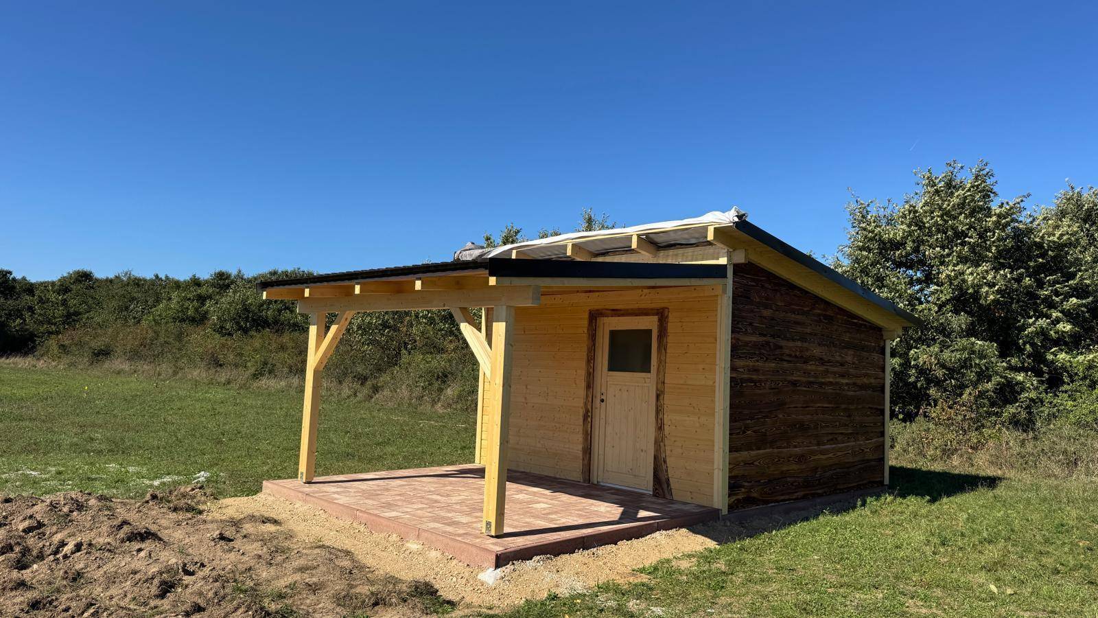 Die Holzhütte von vorne mit einem überdachten Bereich, umgeben von grünen Wiesen.