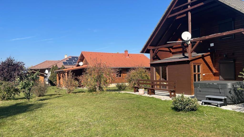 Holzhaus mit Whirlpool und Sitzbank im Gartenbereich unter strahlend blauem Himmel.