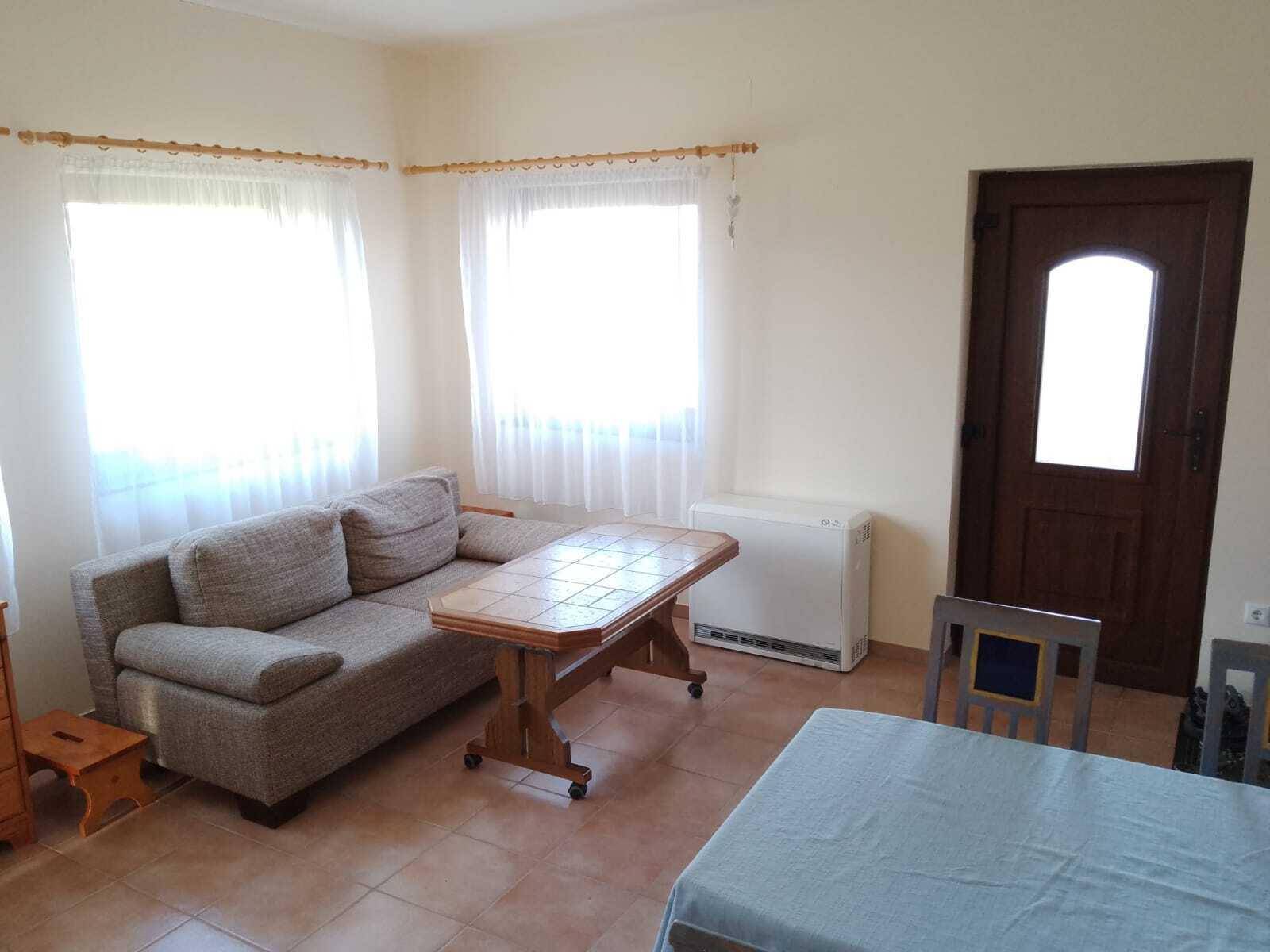 Gemütliches Wohnzimmer mit Sofa, Couchtisch und Fliesenboden, hell durch zwei Fenster.