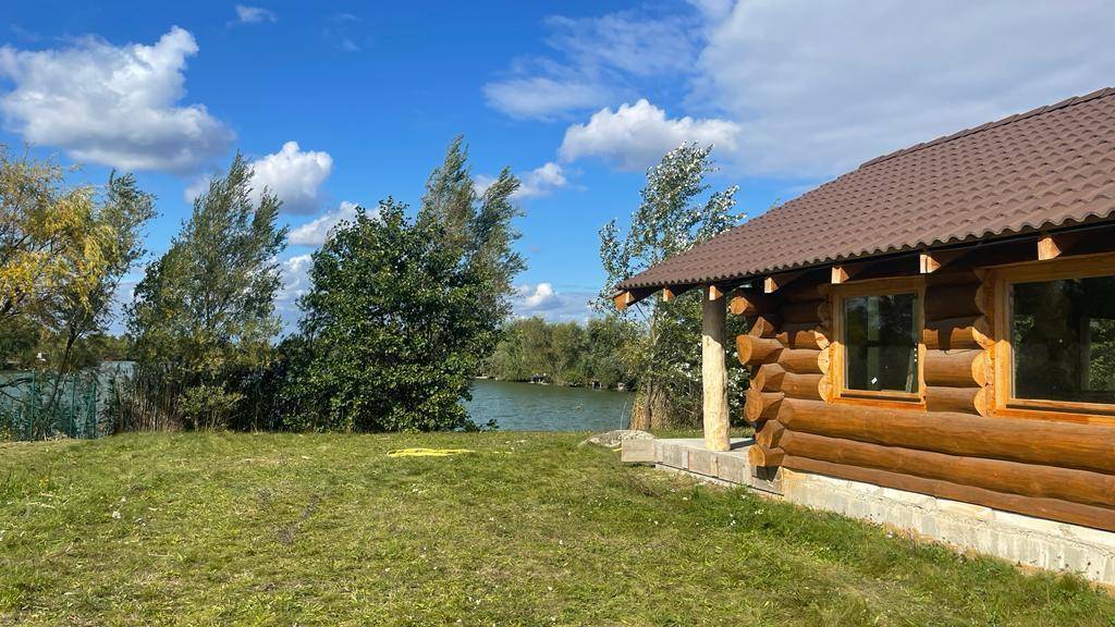 Seitenansicht des Blockhauses mit Blick auf den See und die grüne Uferlandschaft unter blauem Himmel.