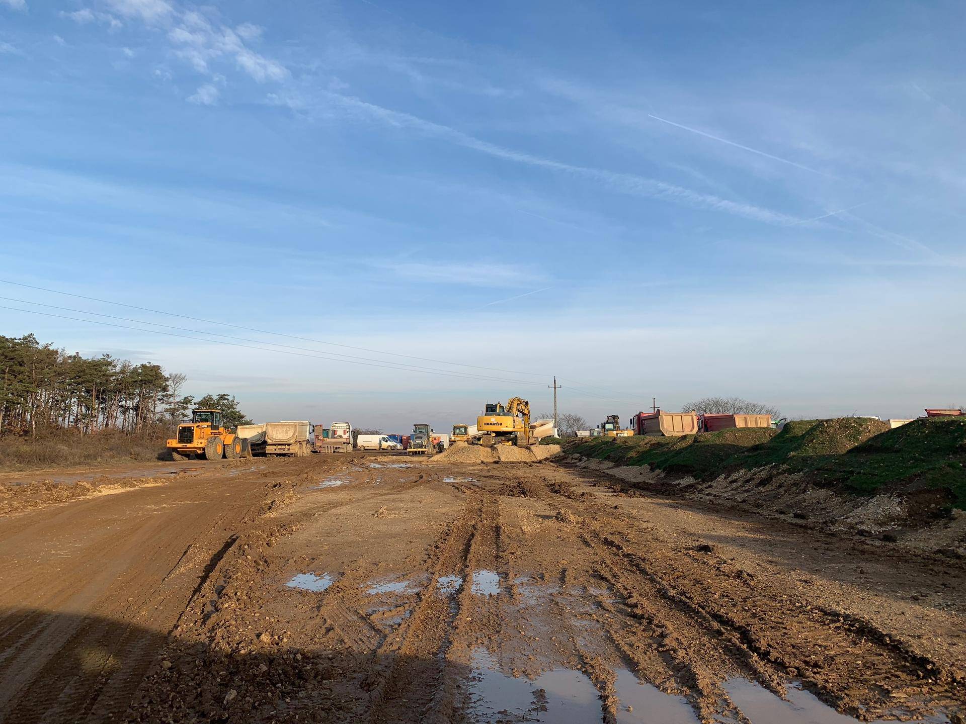 Weitläufige Baustelle mit schlammigem Weg, schweren Maschinen und Erdhügeln unter blauem Himmel.