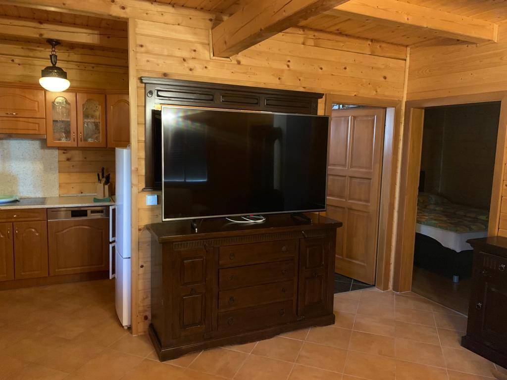 Wohnbereich mit großem Fernseher, Holzmöbeln und angrenzender Küche mit Kühlschrank und Geschirrspüler.