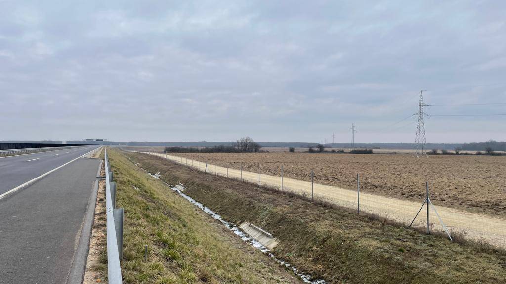 Autobahnabschnitt neben einem weiten Feld, begrenzt durch einen Zaun und Stromleitungen.
