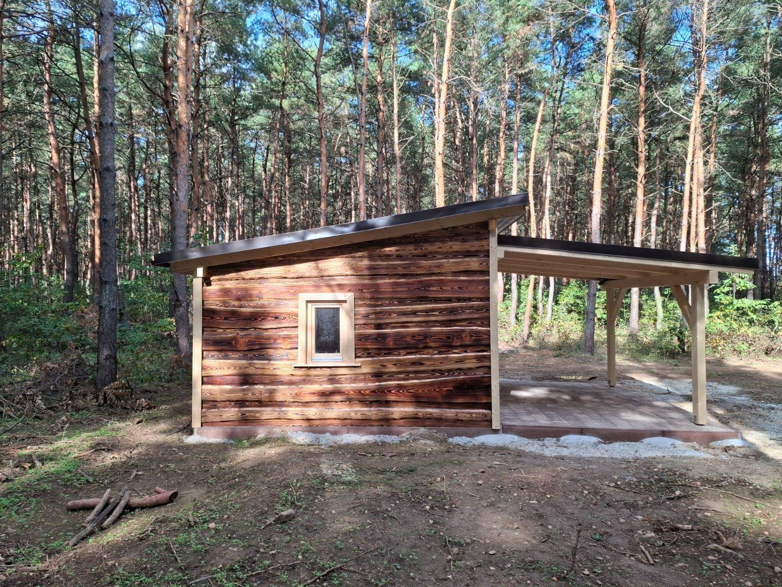 Rustikale Holzhütte mit schrägem Dach und überdachtem Stellplatz, idyllisch im Wald gelegen.