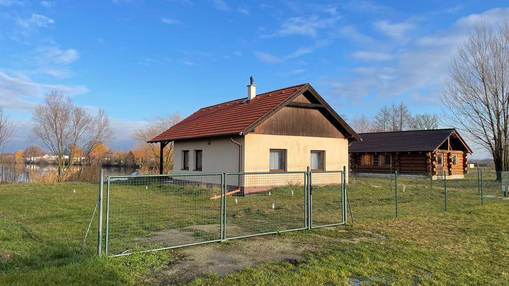 Kleines Haus mit Satteldach und umzäuntem Grundstück, mit See im Hintergrund.