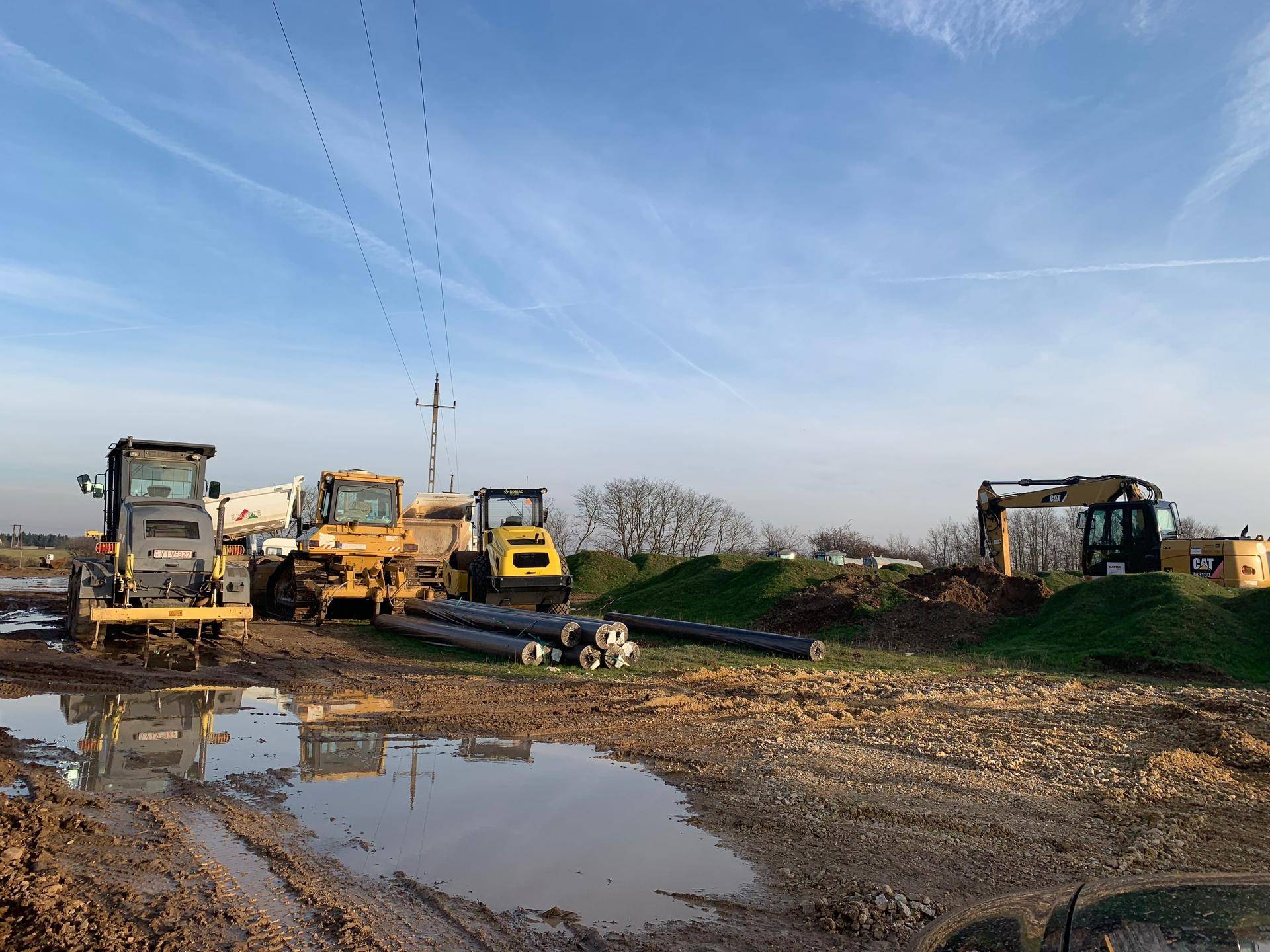 Baustelle mit verschiedenen Baumaschinen, Rohren am Boden und schlammigen Pfützen unter Stromleitungen.