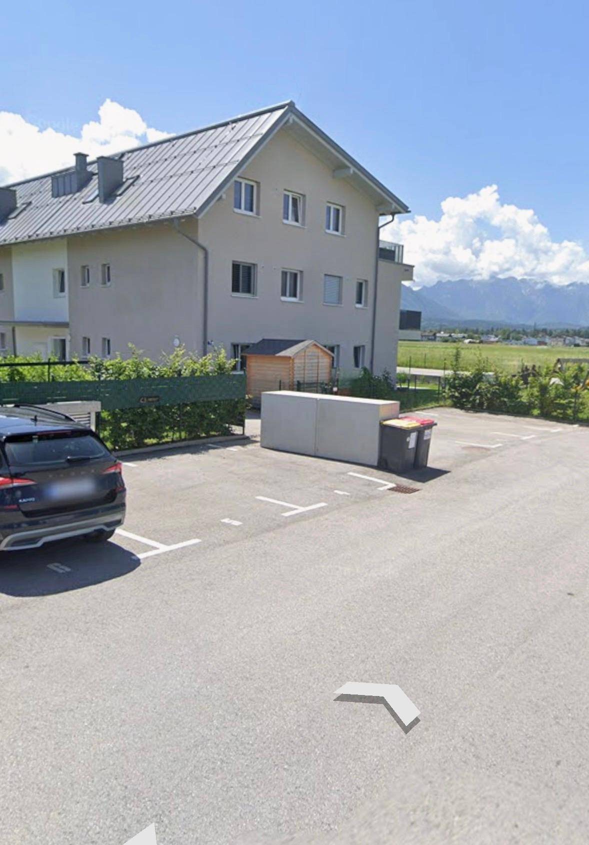 Wohngebäude mit Satteldach und Parkplatz, umgeben von Grünflächen und mit Bergblick.