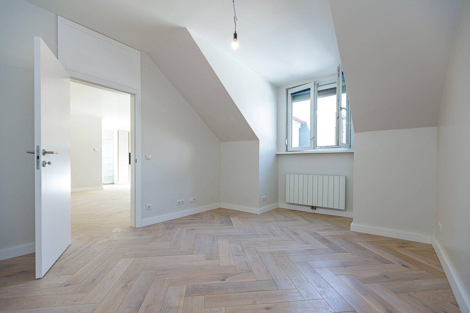 Helles Zimmer im Dachgeschoss mit Fischgrätparkett, schrägen Wänden und Fenster.