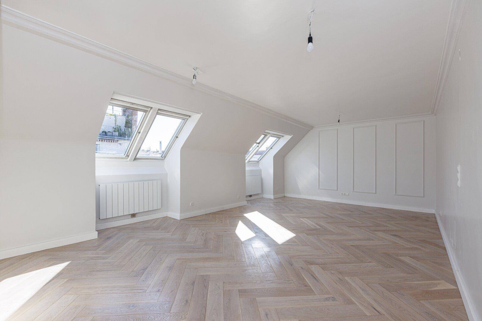 Geräumiges Zimmer mit hellem Fischgrätparkett und schrägen Wänden, durchflutet von Sonnenlicht.