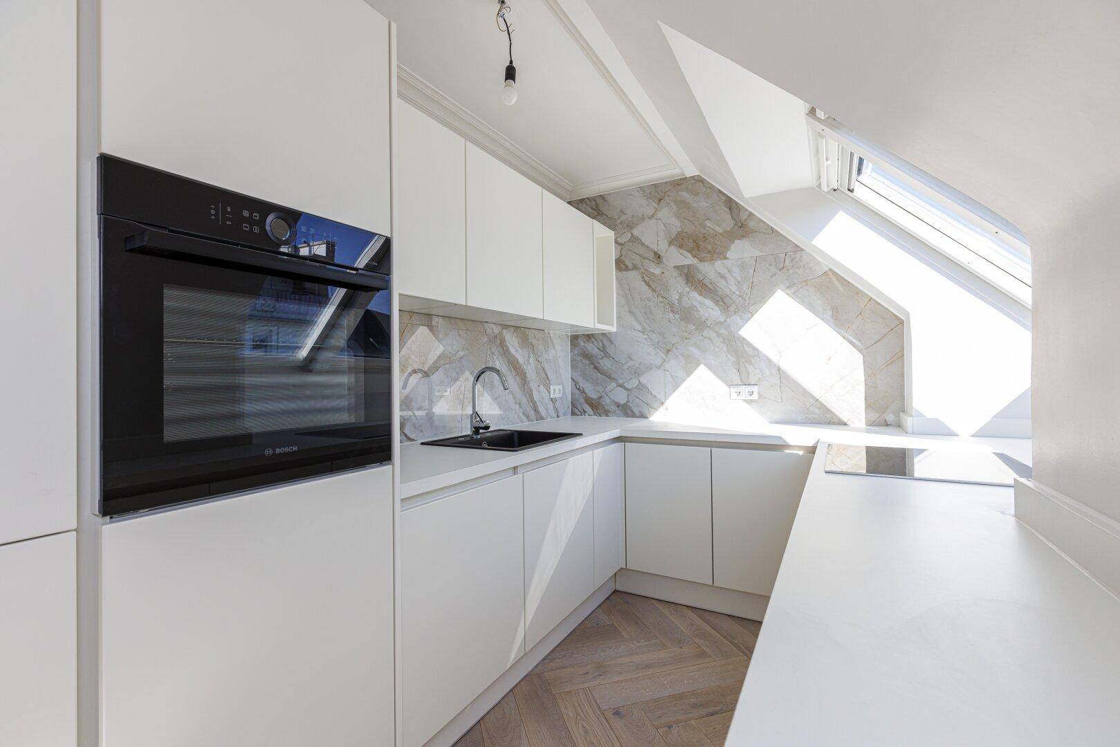 Moderne Küche mit weißen Schränken, integriertem Backofen und stilvoller Steinrückwand unter einem Dachfenster.