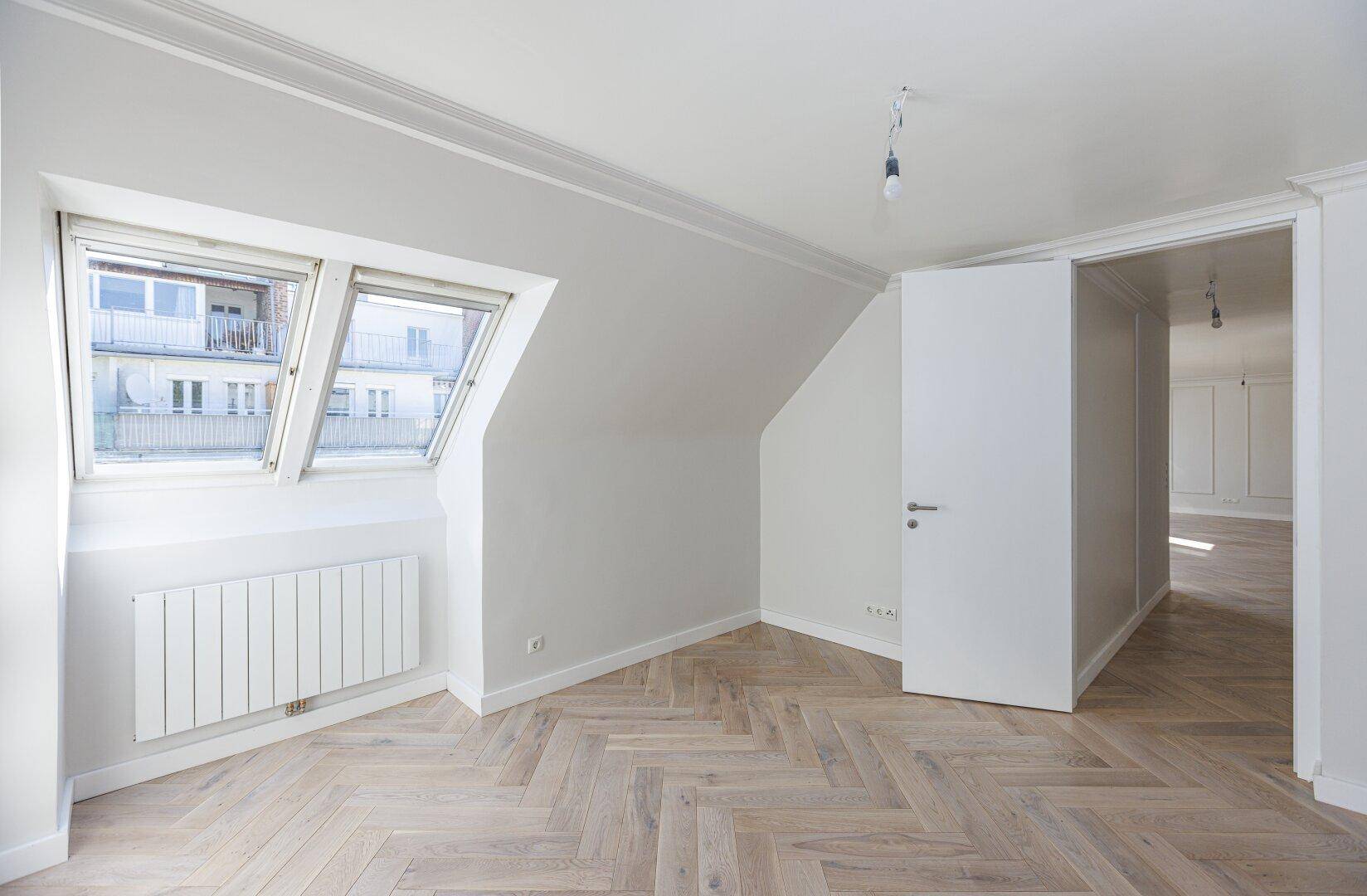 Geräumiges Zimmer mit hellem Fischgrätparkett, Dachfenster und Zugang zu weiteren Räumen.