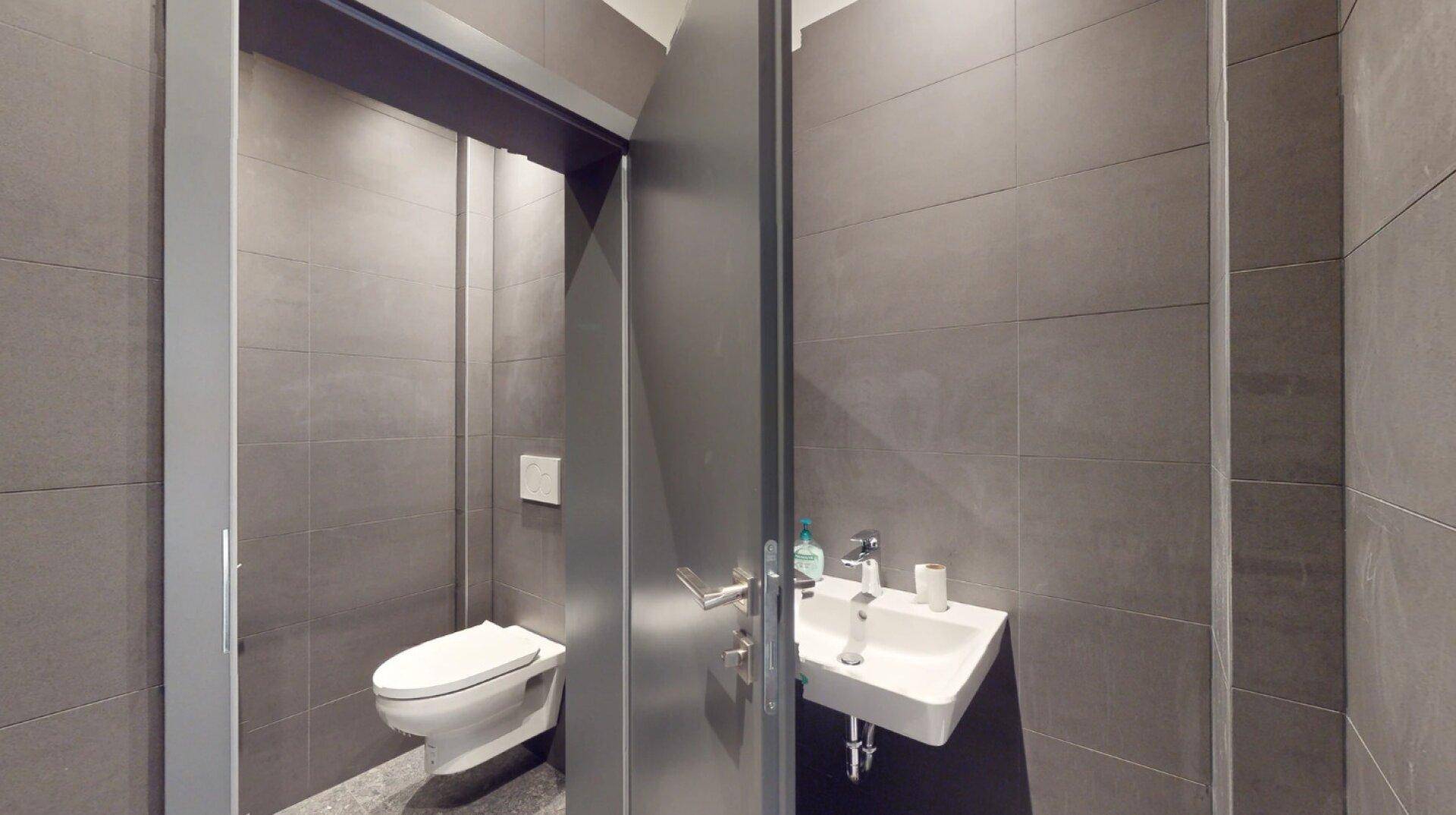 Modernes WC mit grauen Fliesen, wandhängender Toilette und kleinem Waschbecken.