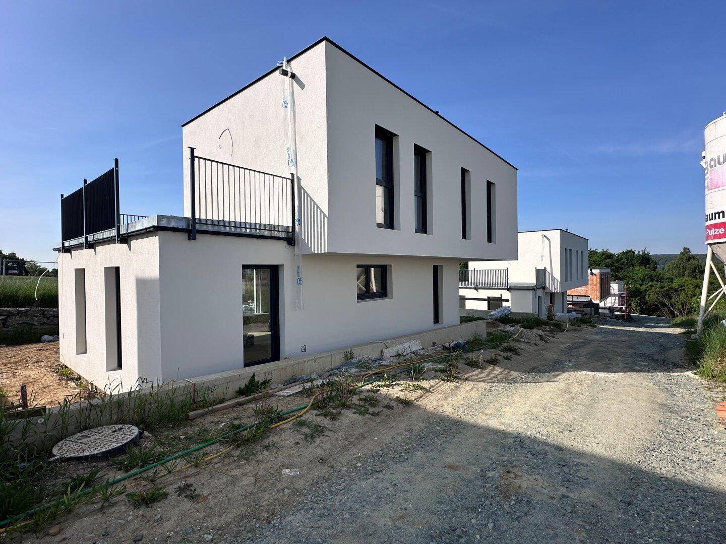 Seitenansicht eines modernen Neubaus mit weißer Fassade und Balkon in sonniger Landschaft.