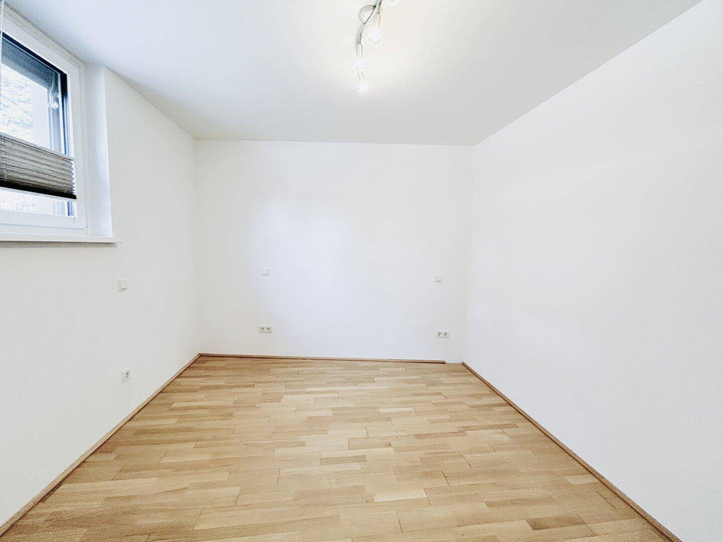 Helles, unmöbliertes Zimmer mit Parkettboden und Fenster mit Jalousie.