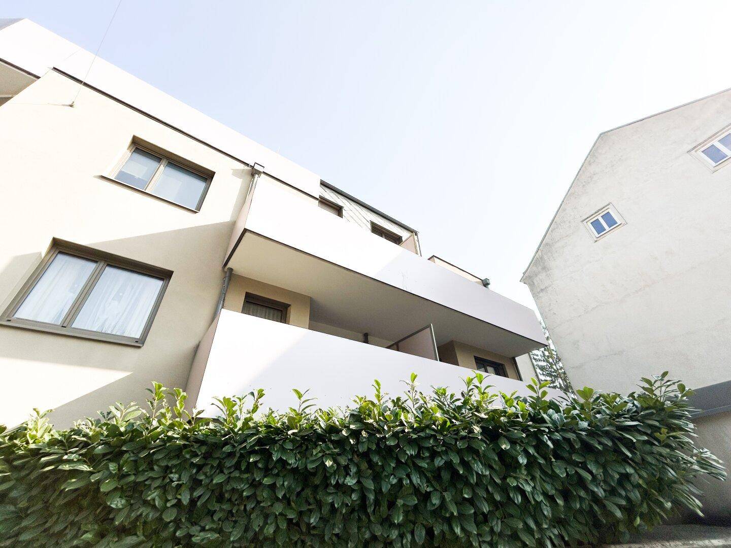 Moderne Gebäudefassade mit Balkonen und grüner Hecke im Vordergrund.