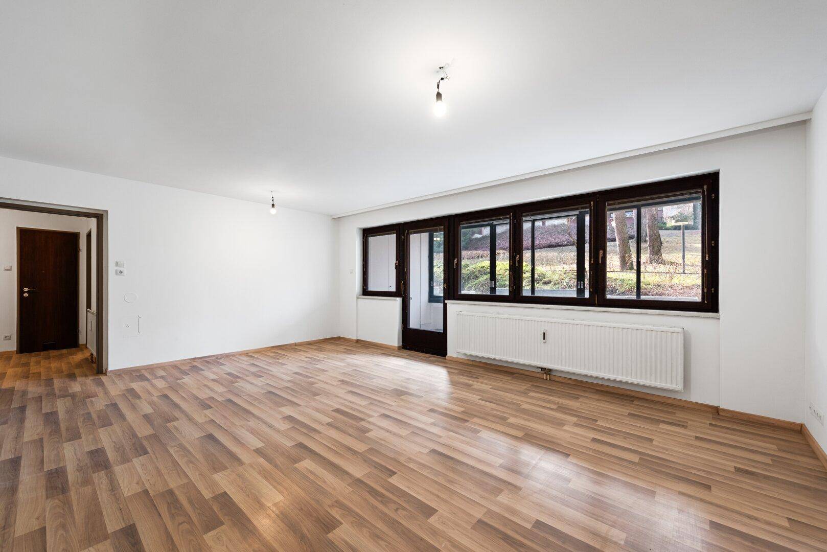 Geräumiges Wohnzimmer mit Laminatboden, weiß gestrichenen Wänden und Zugang zu einem weiteren Raum.