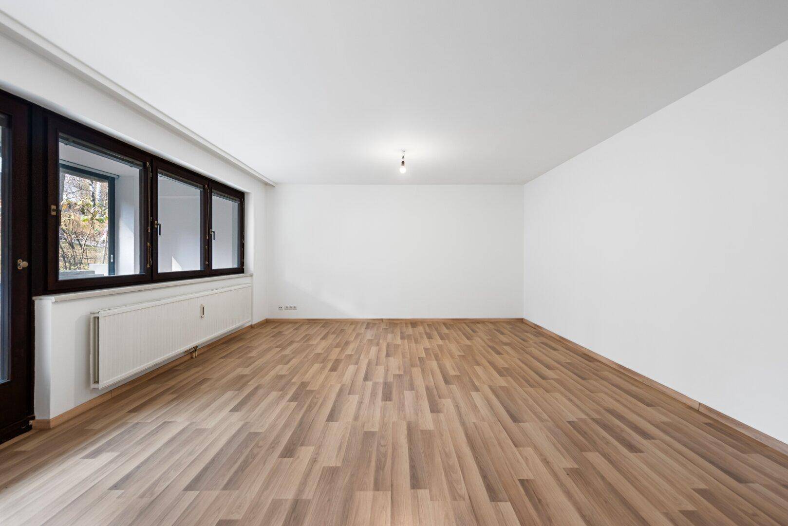 Leeres Wohnzimmer mit Laminatboden und großen Fenstern, die eine helle und offene Atmosphäre schaffen.