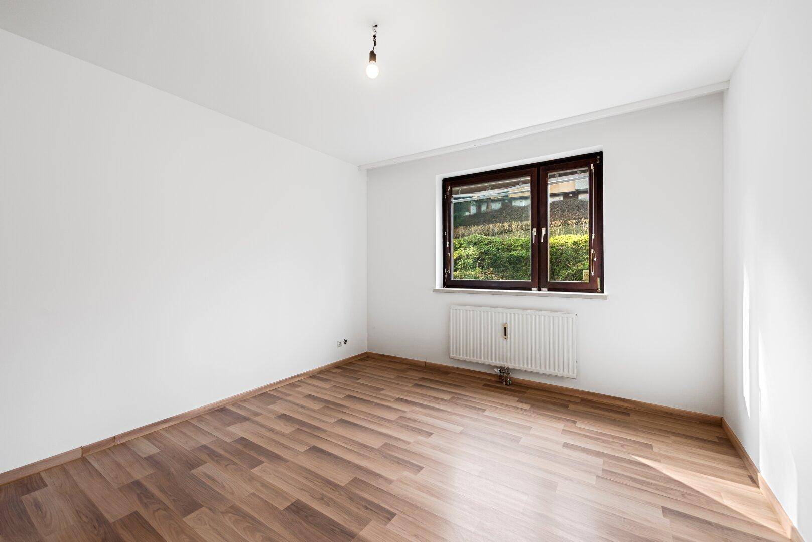 Leeres Zimmer mit Laminatboden und Fenster, das viel Tageslicht hereinlässt und einen Blick ins Grüne bietet.