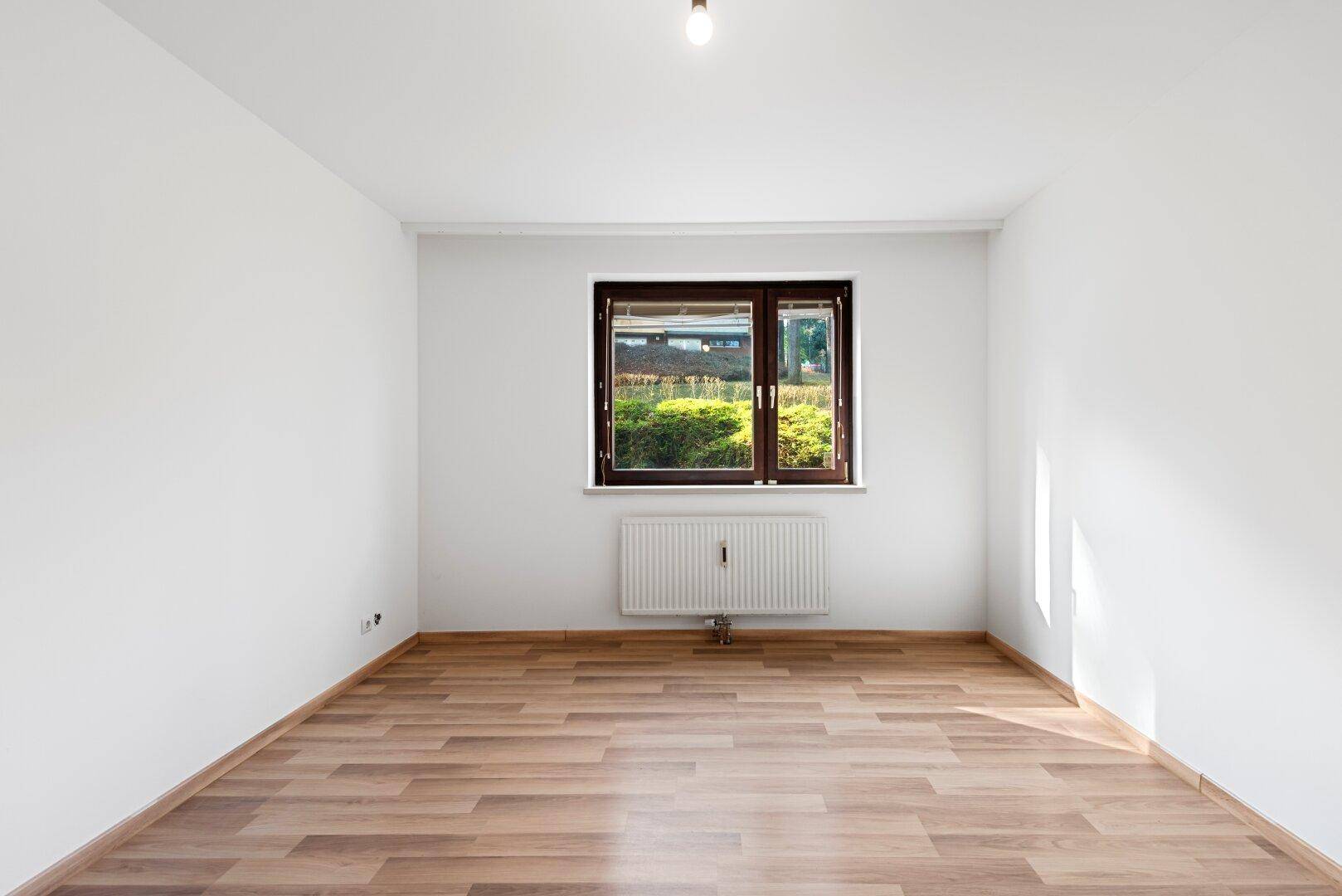 Helles Zimmer mit Laminatboden und einem Fenster, das den Blick auf die umliegende Natur freigibt.