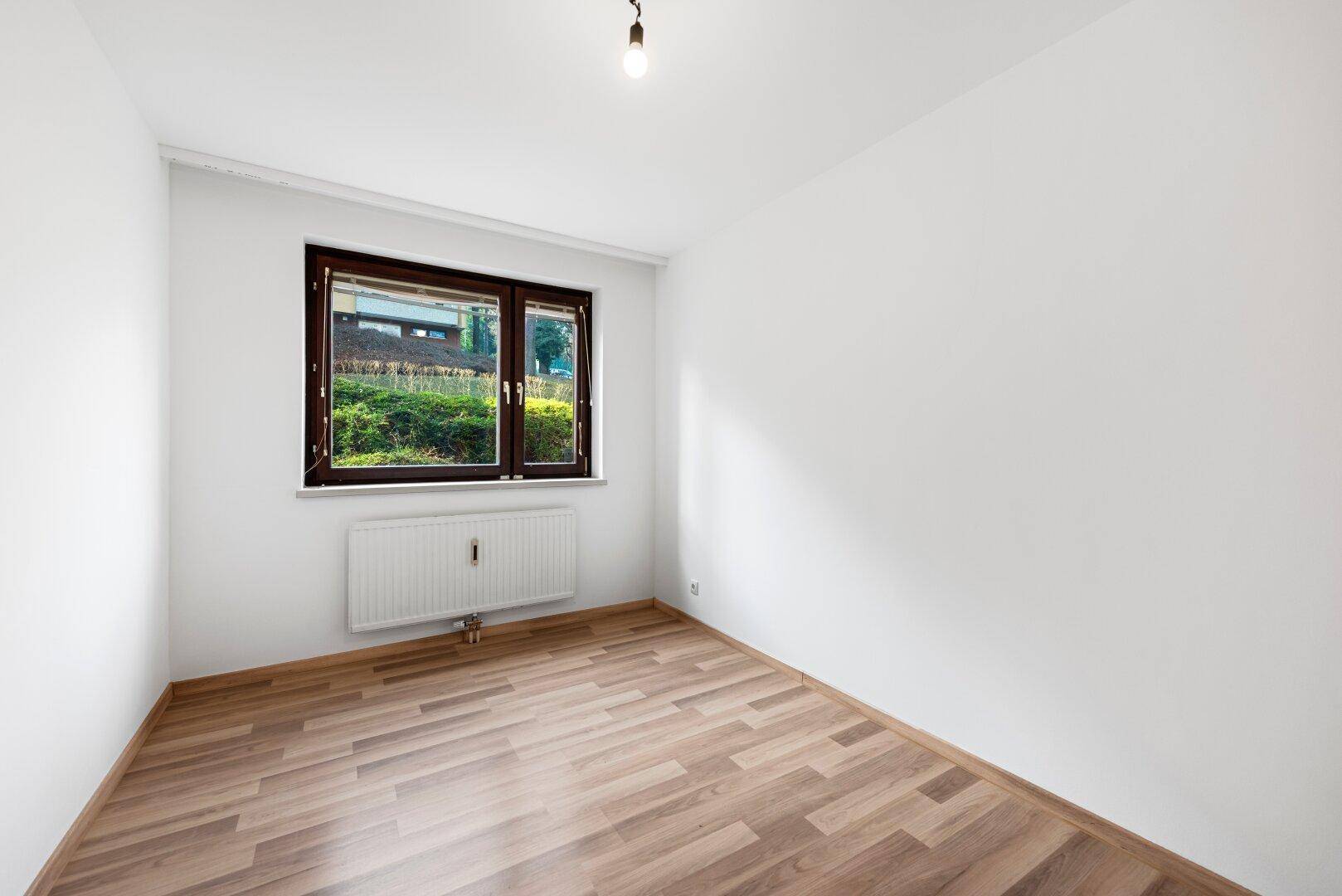 Geräumiges Zimmer mit Laminatboden und Fenster, das eine angenehme Helligkeit und einen grünen Ausblick bietet.