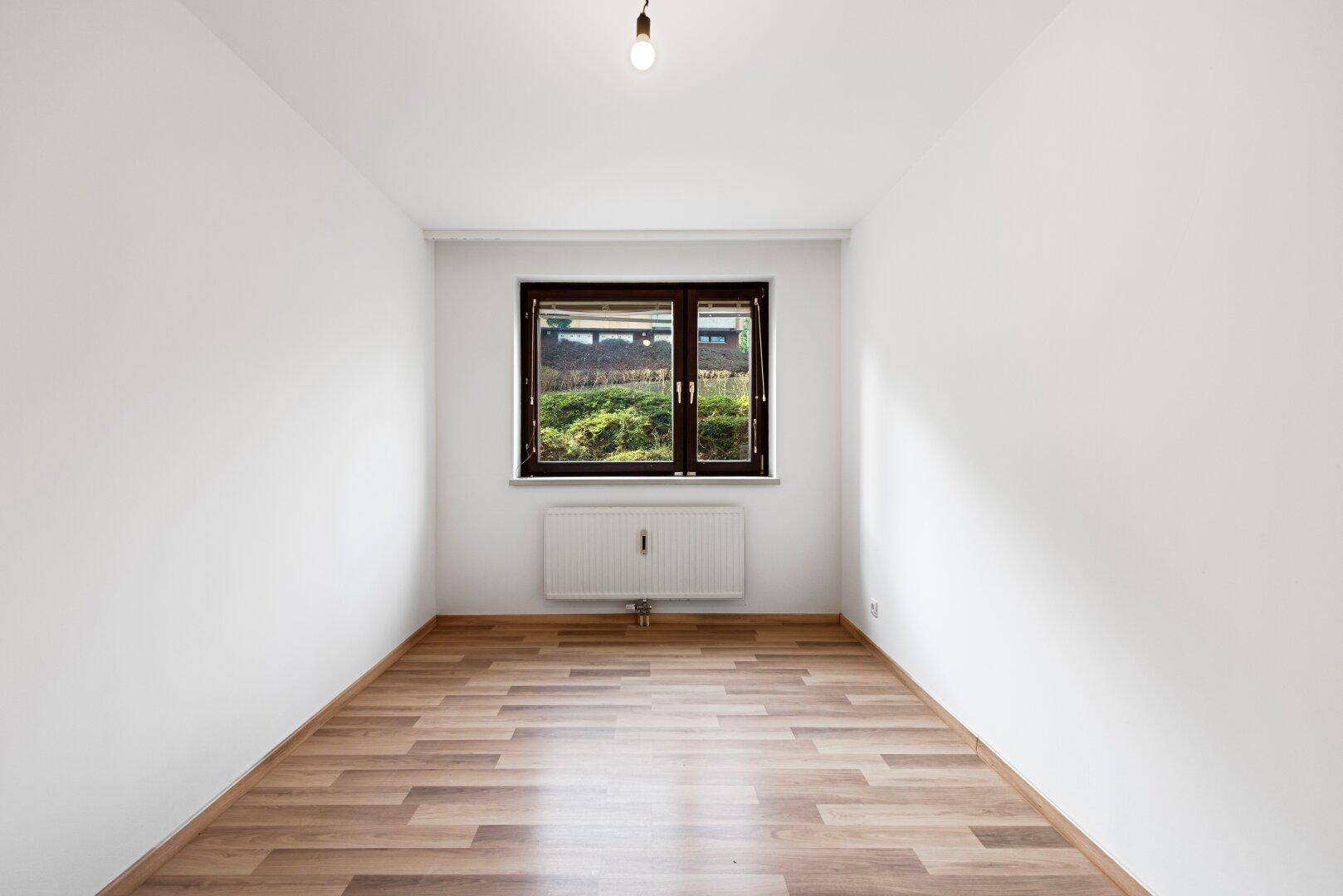 Leeres Zimmer mit Laminatboden und Fenster, das den Raum mit natürlichem Licht durchflutet.