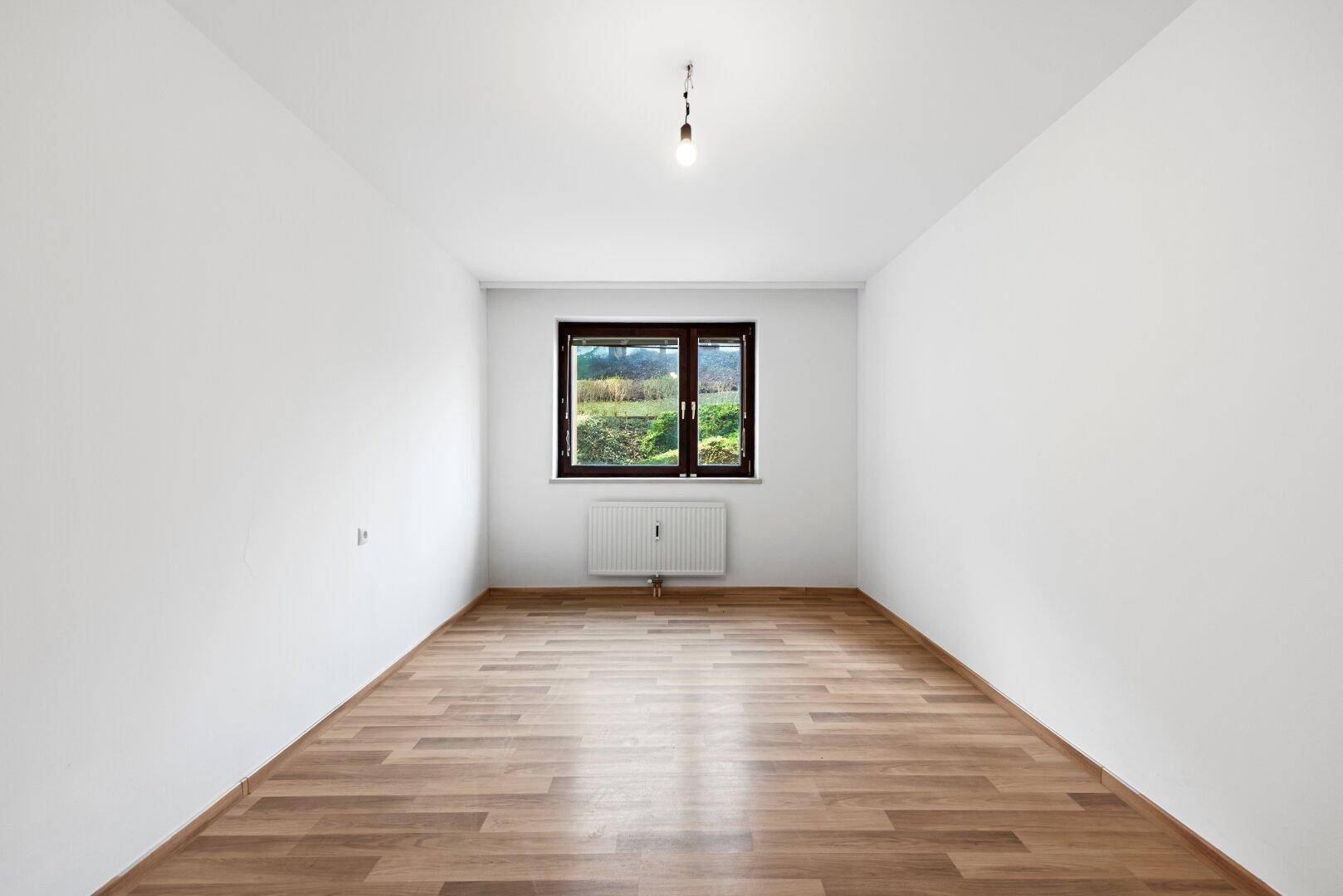 Helles Zimmer mit Laminatboden und Fenster, das einen ruhigen Blick ins Grüne ermöglicht.