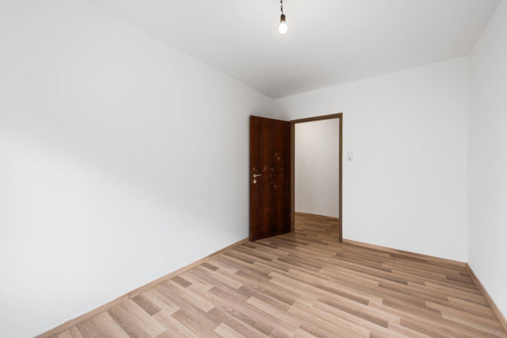 Leeres Zimmer mit Laminatboden und offener Holztür, die den Zugang zum Flur ermöglicht.