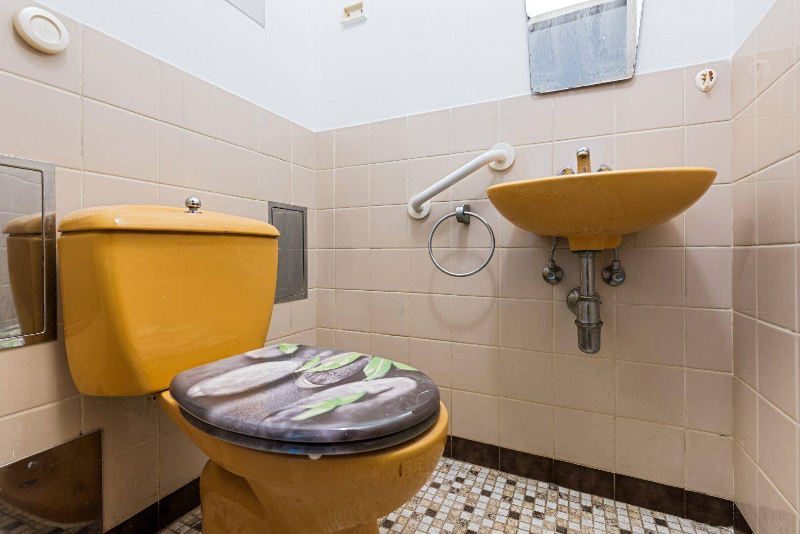 Separate Toilette im 80er-Jahre-Stil mit gelber Keramik und Fliesenboden, inklusive Haltegriff.