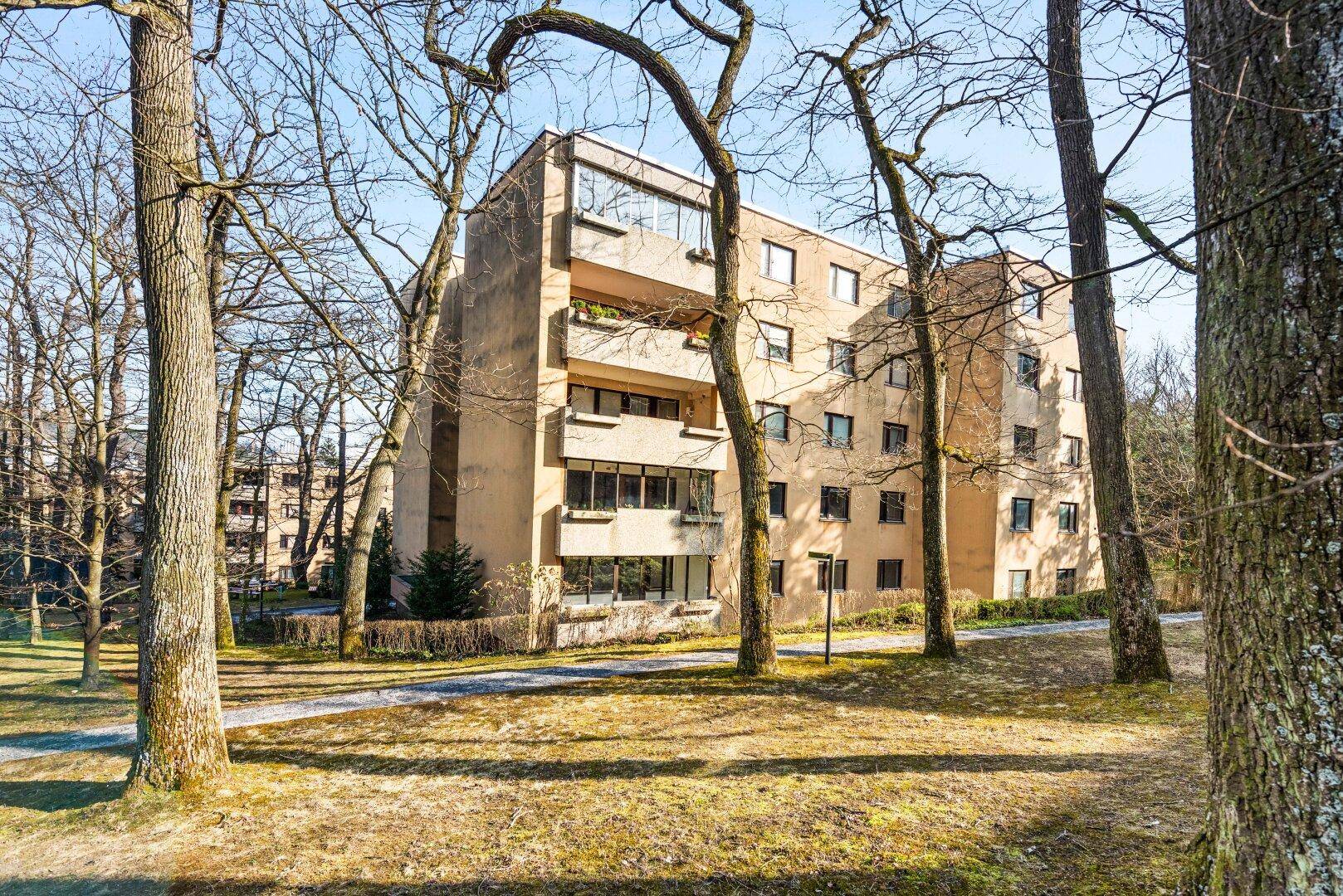 Mehrstöckiges Wohngebäude umgeben von Bäumen und Grünflächen an einem sonnigen Tag.